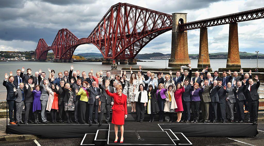 Nicola Sturgeon, in a red blazer and dress, stands with the 56 SNP MPs who won seats in the 2015 election. They are in front of the Forth Road Bridge.