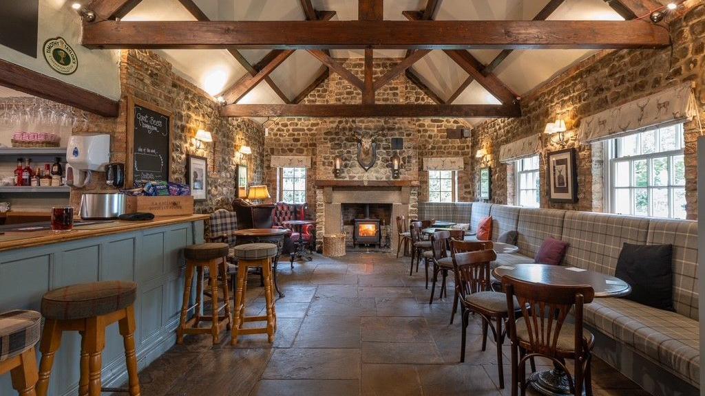 The interior of a country pub, with brown tiled flooring and wooden ceiling beams. There is a blue-painted bar with wooden bar stools on one side, and grey booths with small circular tables on the other side, and a wood burner at the far end.