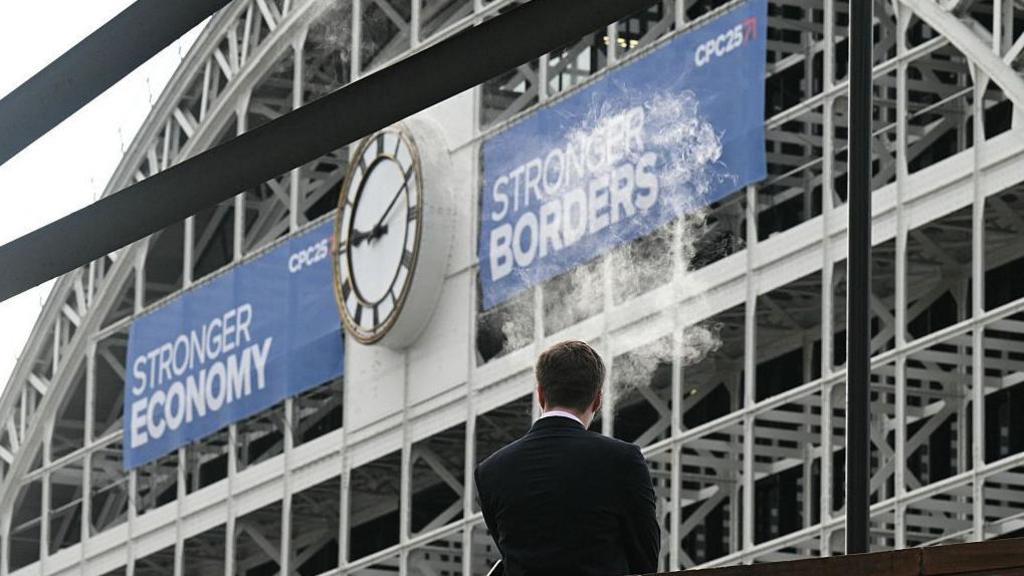 A man vapes into the air outside the venue on the third day of the annual Conservative Party conference in Manchester. He is standing in front of the former railway station and large blue banners saying Stronger Economy and Stronger Borders are hanging either side of the large station clock.  