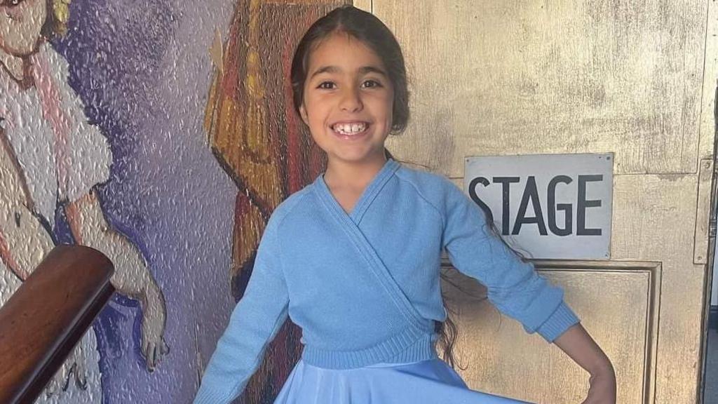 Image shows Alice Aguiar, wearing a blue cardigan and blue ballet costume. She is standing in front of a sign painted on a door. The sign says 'stage'