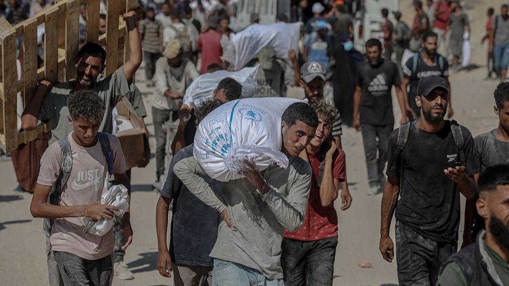 Group of people walk on grey sand. A number struggling to carry large bags of food on their shoulders.