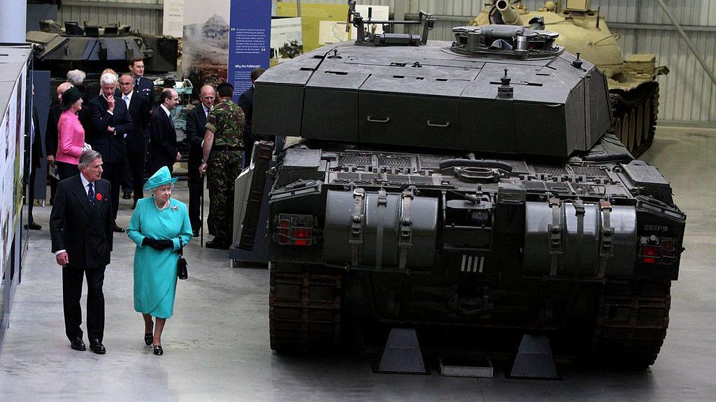 The then director, Sir Roger Wheeler shows Queen Elizabeth II around the museum. He wears a dark shut and a red poppy. Her Majesty is dressed in a blue dress and hat. They are walking next to a large tank. 