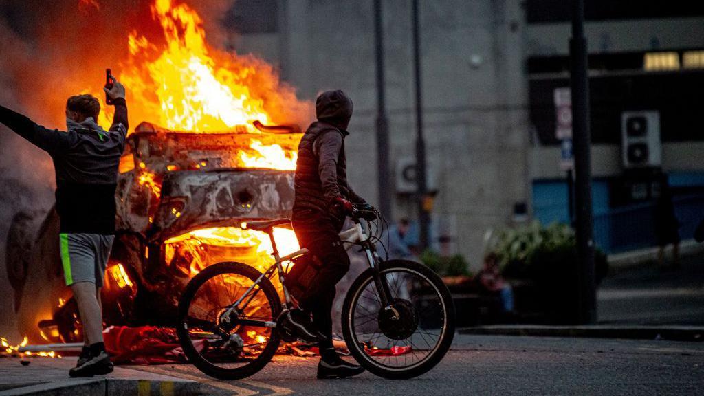 An image of a riot in Sunderland shows two young people, one wearing shorts with his arms in the air and another in a gilet and hoodie on a bicycle. They are standing in front of a burning vehicle