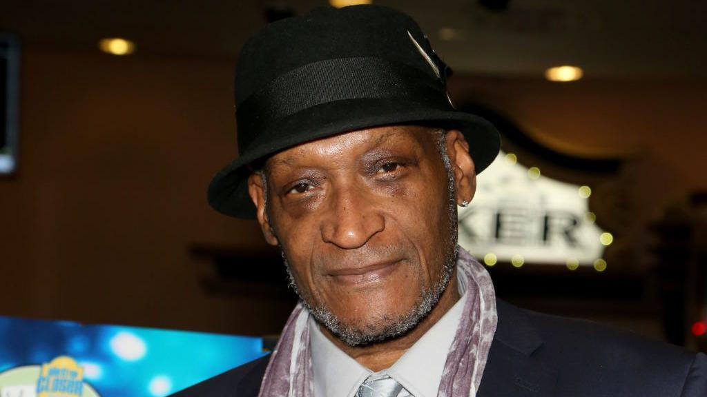 Tony Todd, looking directly to camera. He is wearing a black bowler hat and has light, grey stubble around his jawline