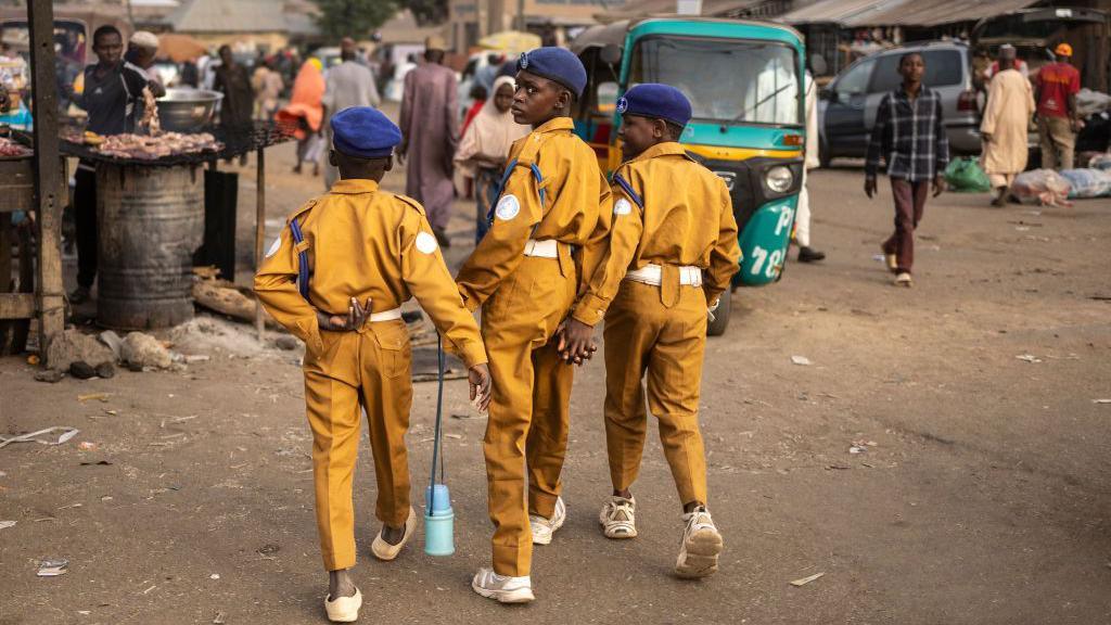 Three boys walk away from the camera, wearing yellow trousers and shirts with a blue hats. Market traders sell goods nearby in Jos, Nigeria - Thursday 27 February 2025.