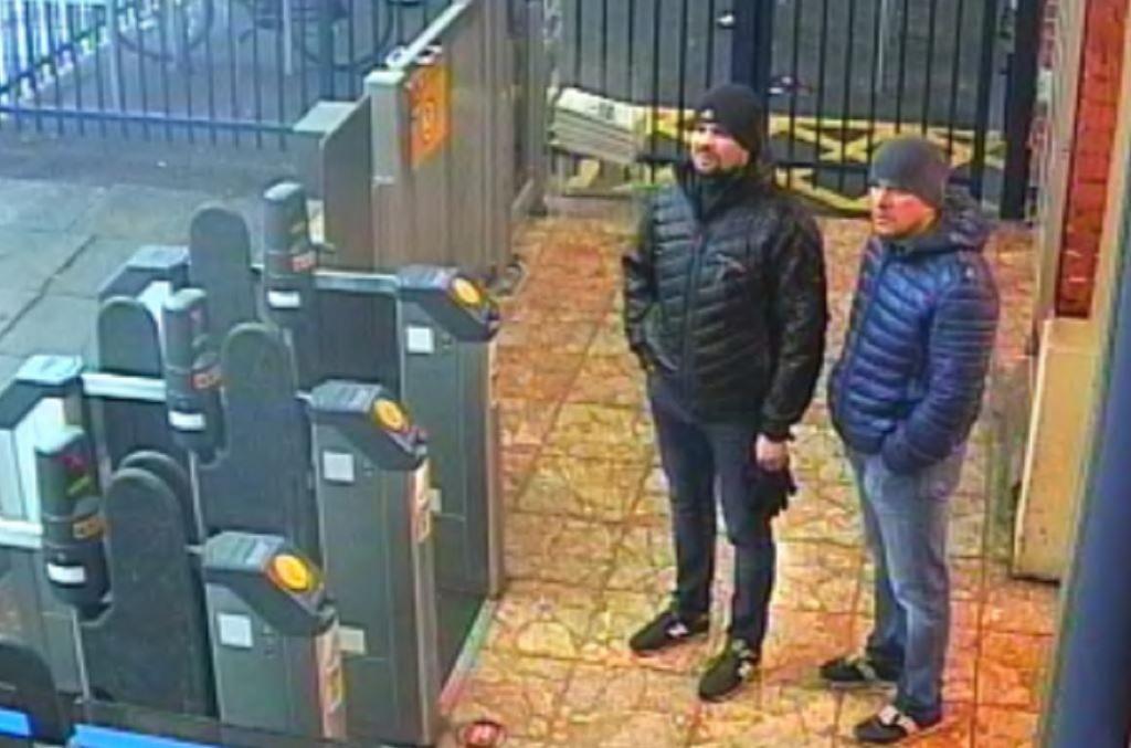 A bearded man in a black beanie hat, black puffa jacket, blue jeans and black trainers stands next to a man in a grey beanie hat wearing a blue puffa jacket with blue jeans and trainers close to ticket barriers in a London Underground station.