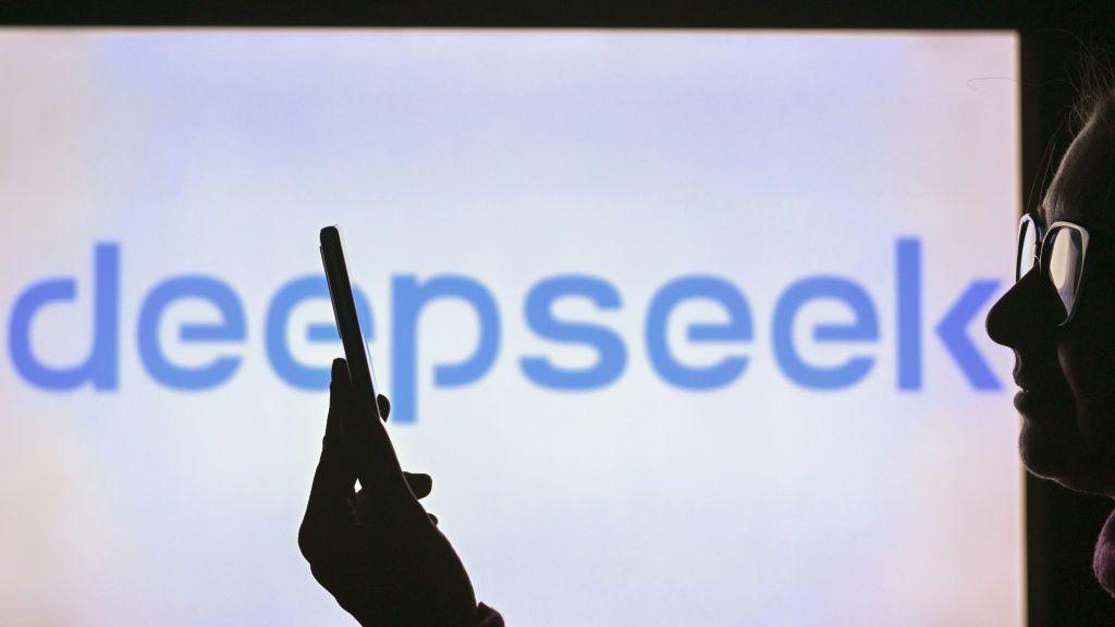 A woman holds a cell phone in front of a computer screen displaying the DeepSeek logo