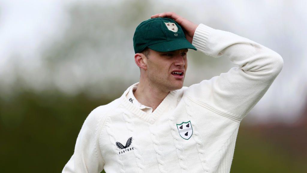 Worcestershire bowler Ben Gibbon puts his left hand on top of his green club cap as he looks at the action while in the field