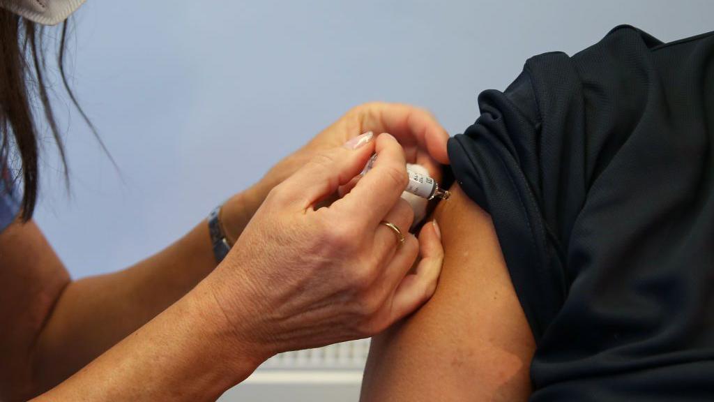 Pictured is a person's arm, with their sleeve rolled up, with a medic's hands administering a vaccination