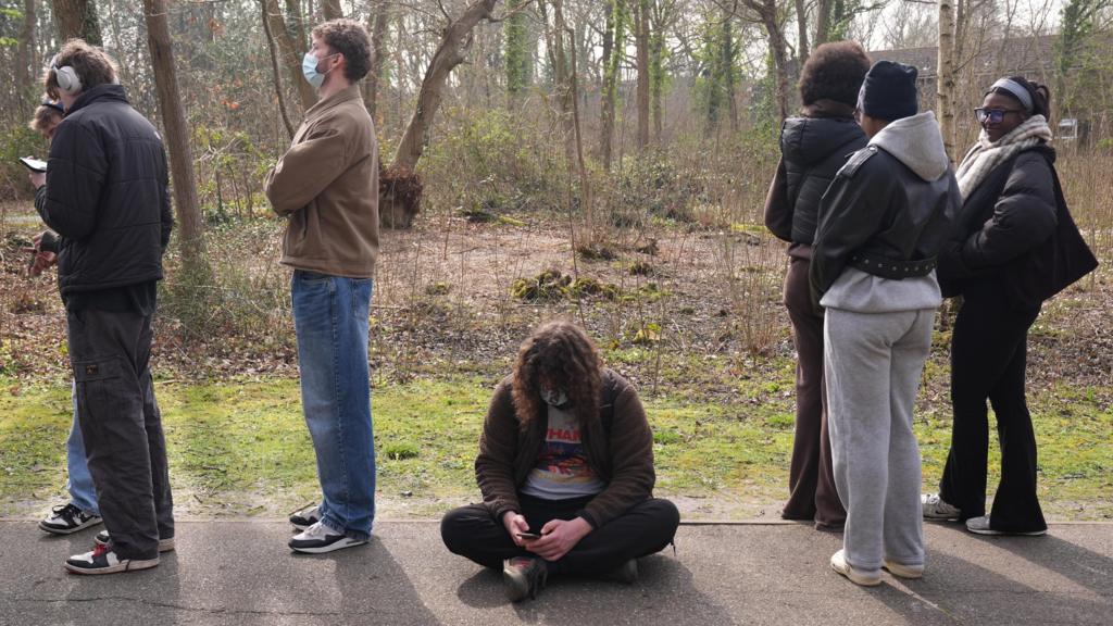 A person sits on the floor in a mask as people queue around them