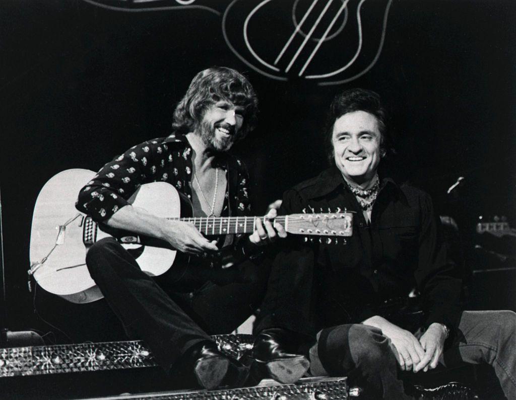 Kris Kristofferson, holding a guitar, poses with country music legend Johnny Cash on the set of a TV show in the 1960s.