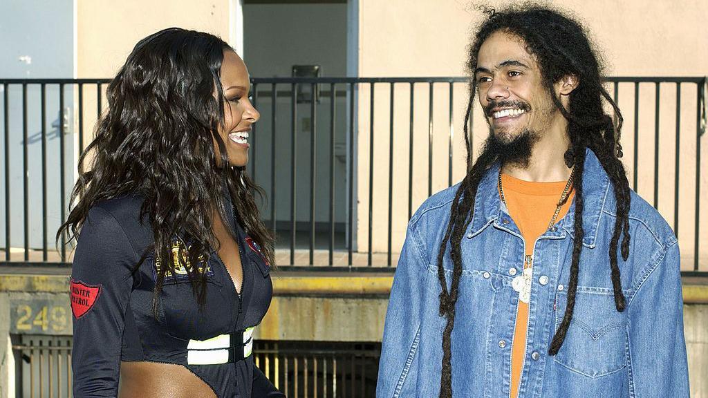 Samantha, left, with long curly brown hair, wearing a navy bodysuit with cutaway details. She is stood outside, against a railing, smiling at Damian Marley who is stood beside her. He has long dreadlocks and a goatee, wearing a denim jacket and orange top. He is smiling widely.