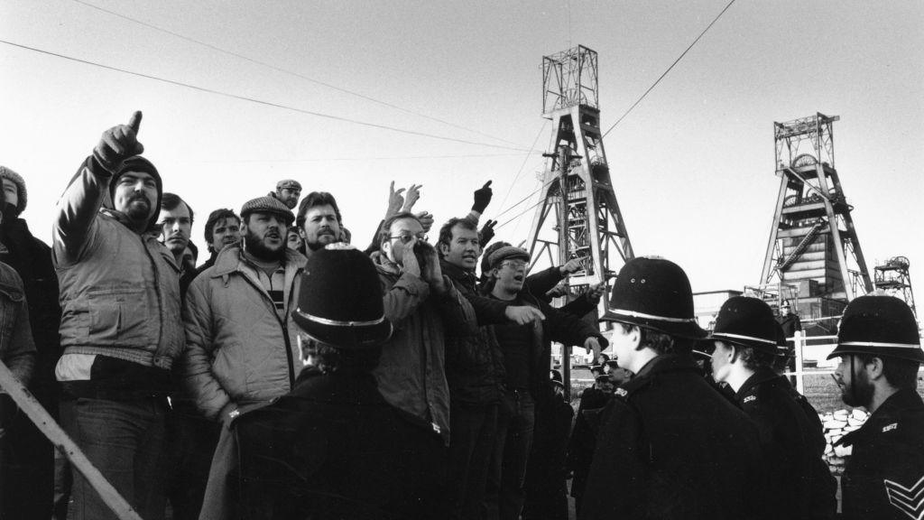 Men stand behind a police line. They are shouting and gesticulating with a mine in the background. 