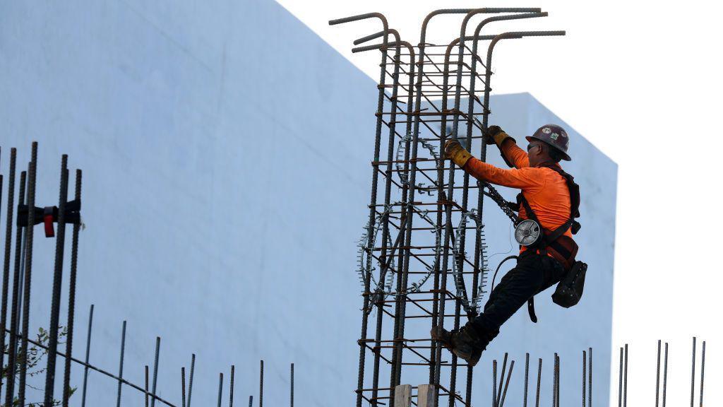 A construction worker in an orange shirt and wearing a hard top hat helps build a support column using steel rebar during the building of a condo tower on February 10, 2025 in Miami, Florida. 