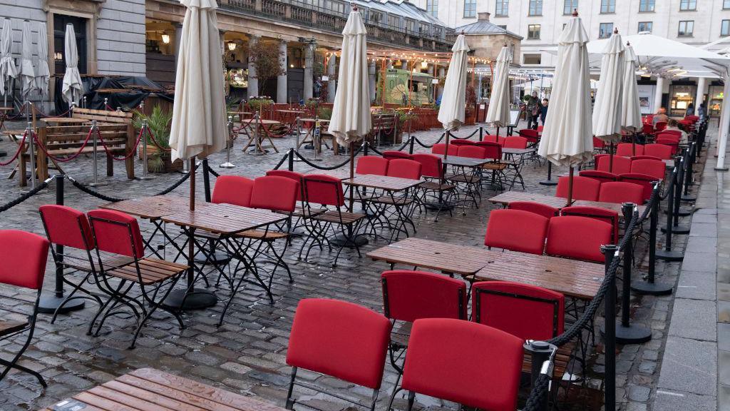 Rows of empty wooden tables and red chairs arranged for outdoor dining in a cobblestoned courtyard, with closed white parasols and roped-off sections, in front of a building with arched windows and covered walkways.