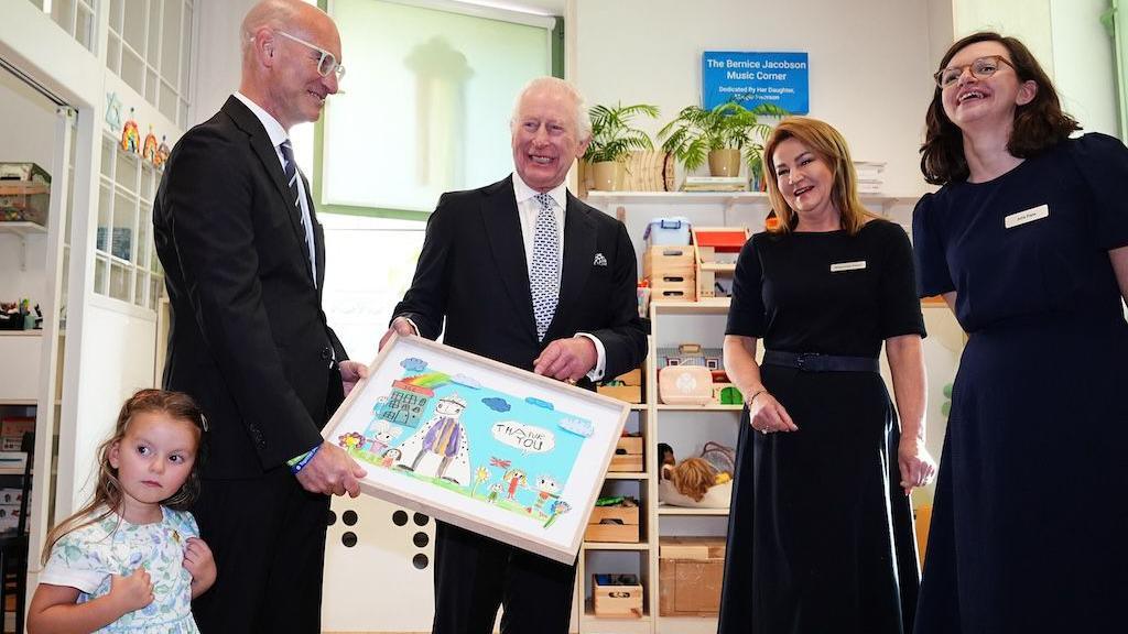 King Charles III being presented with a painting. He is smiling for a photo with a man, two women and a young girl.