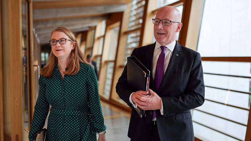 Shirley-Anne Somerville - a woman with red shoulder length hair in a green dress - and first minister John Swinney - a bald man in a dark suit with purple tie - walking together into the Scottish Parliament chamber