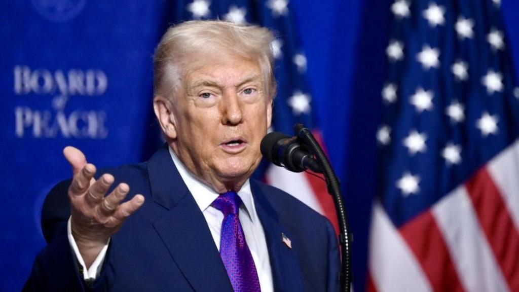 Donald Trump, wearing a purple tie, white shirt and blue suit gestures in front of a blue backdrop and an American flag