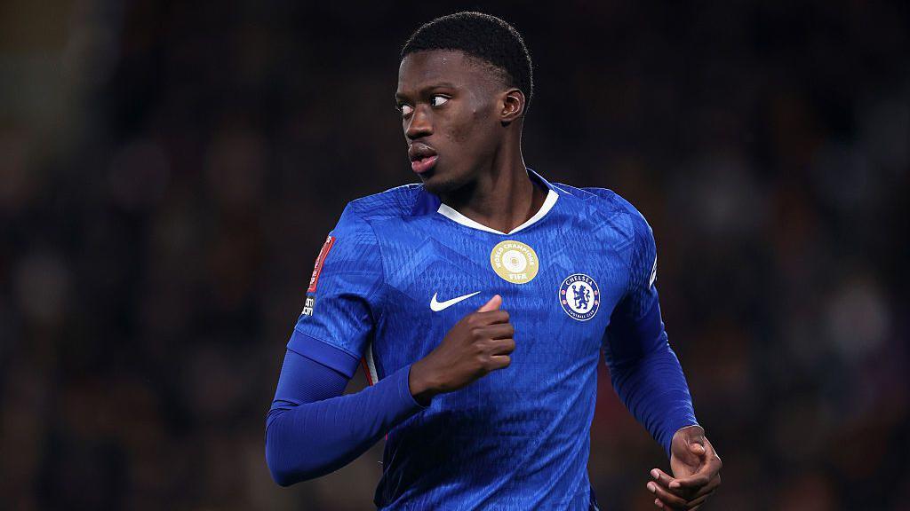 Mamdou Sarr visible from waist up running on the pitch against Hull City. He is wearing a blue Chelsea home jersey with matching blue undersleeves and is turning his head to look to his right