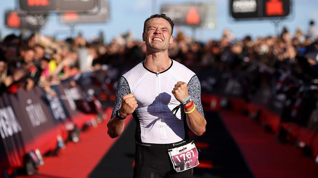 Lewis Eccleston wears a white and block top as he looks up in delight at the finish line