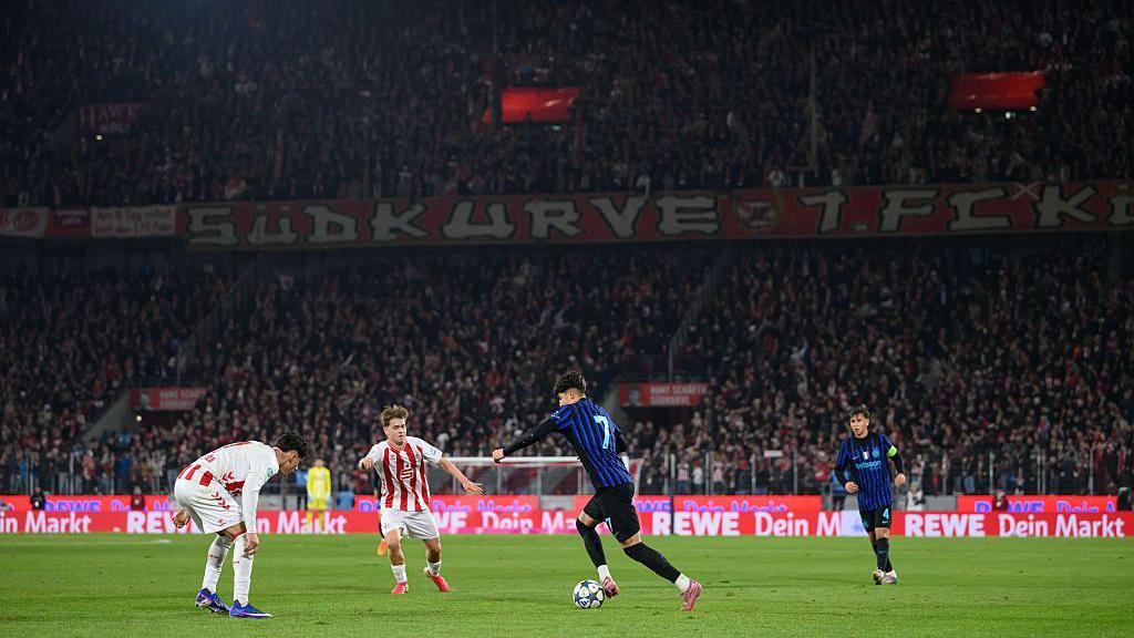 Inter Milan's number seven runs with the ball as two Cologne players close him down. Thousands of fans fill two tiers of the ground around the pitch.