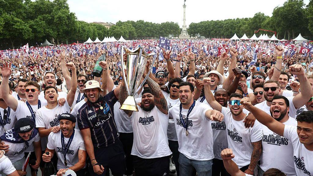 Bordeaux Begles players celebrate last year's Champions Cup victory at an event in the city, lifting the large trophy as a flag-waving crowd stand behind them. The players wear white T-shirts with the slogan WE ARE CHAMPIONS