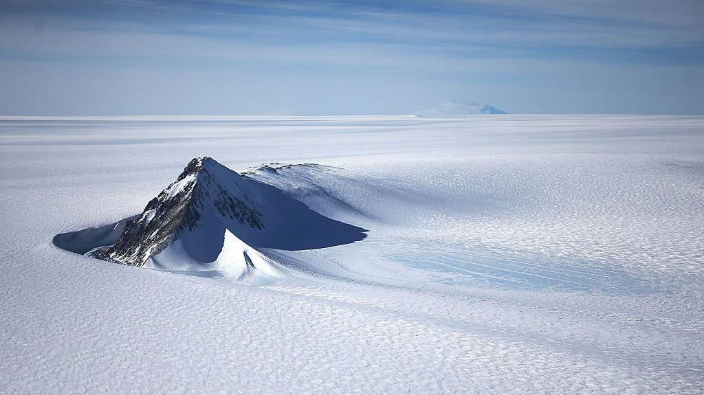 A section of the West Antarctic Ice Sheet with mountain peaks poking out of the pristine, flat, white icy surface. The sky above is light blue with light clouds.