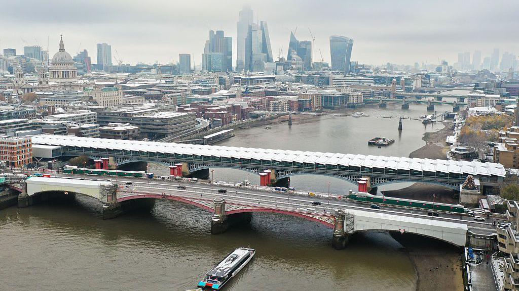 An aerial view shows snow-covered buildings and offices, backdropped by skyscrapers in the City of London, as an Uber taxi boat passes under crossing under Blackfriars Bridge on the River Thames.
