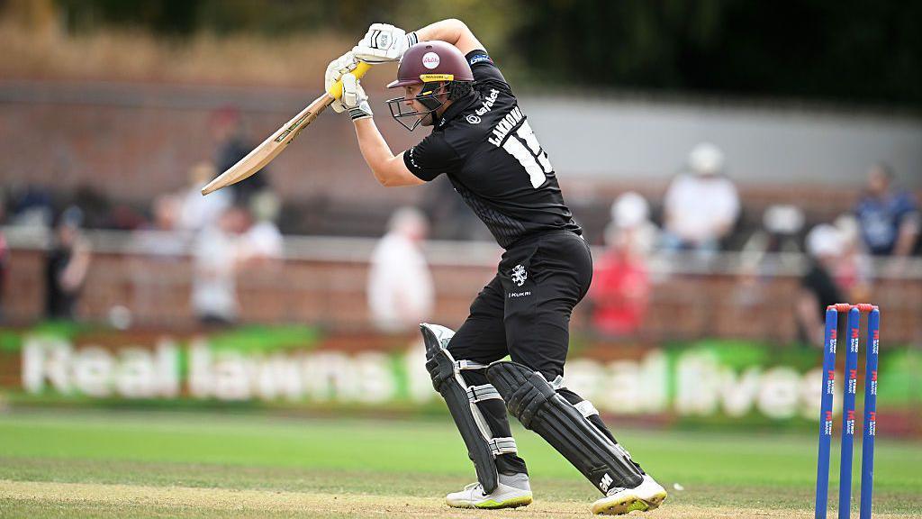 A man in cricket gear is on a cricket pitch swinging a bat. He is captured in motion with his arms above his head.
