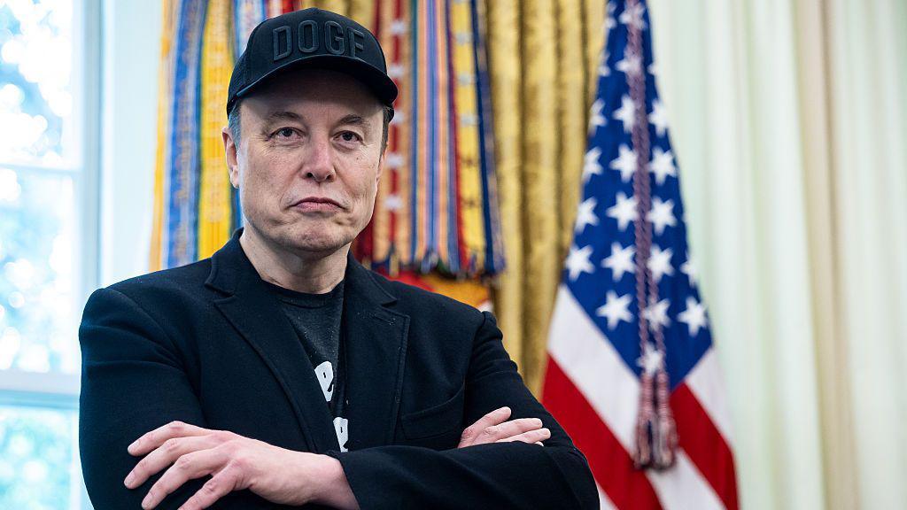 Elon Musk in the Oval Office. He is wearing a dark blazer and a baseball cap. In the background a US flag is visible. 