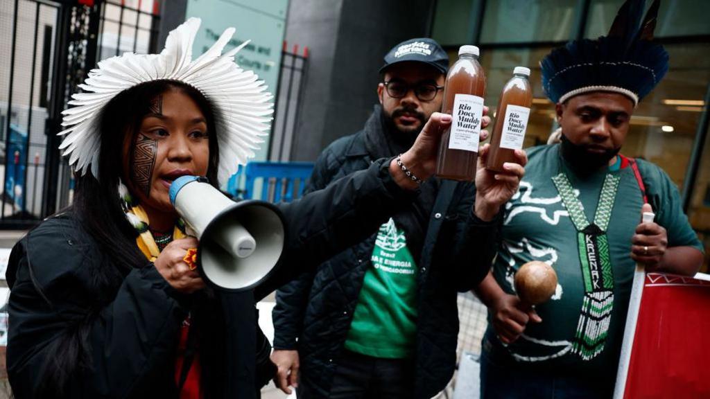 Wakrewa Krenak, left, who lives in Conselheiro Pena, 400km from Mariana, holds a bottle of the contaminated water retrieved from the river Doce, as she speaks in a loud speaker during a victim gathering outside the High Court in London, on 21 October 2024.