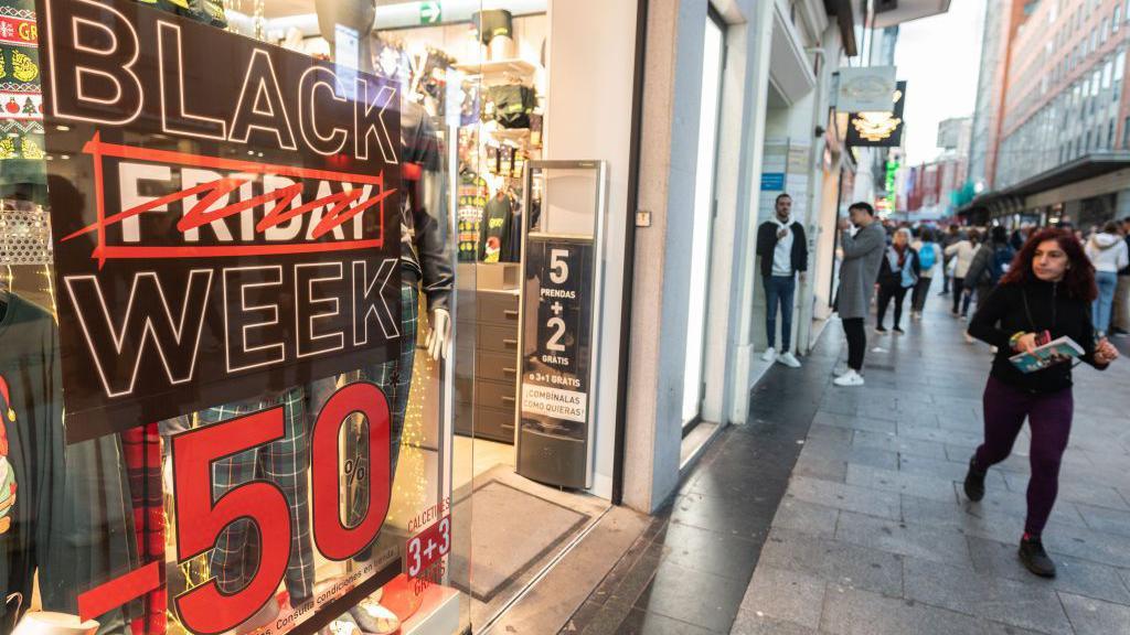 A woman walks past a sign on the high street that says Black Friday Week - 50% and the work Friday is crossed out