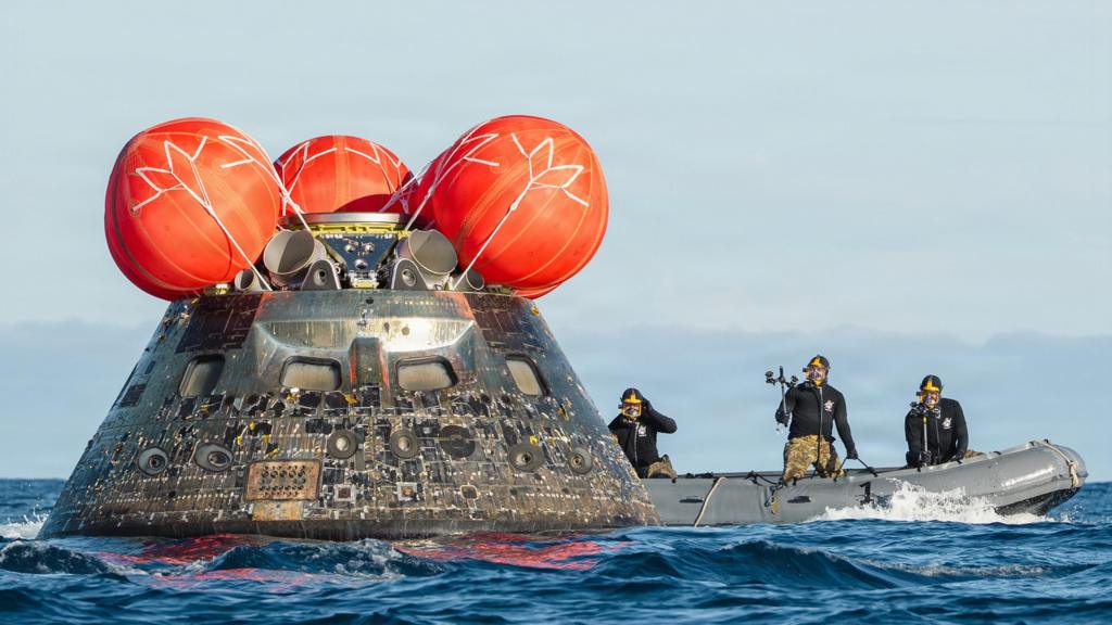 The artemis crew's capsule in the ocean with a rubber boat nearby with three people in wetsuits