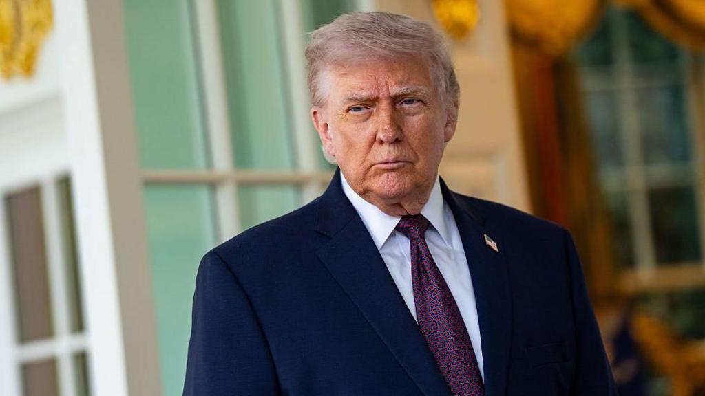 Donald Trump stands outside the Oval Office in a navy suit.