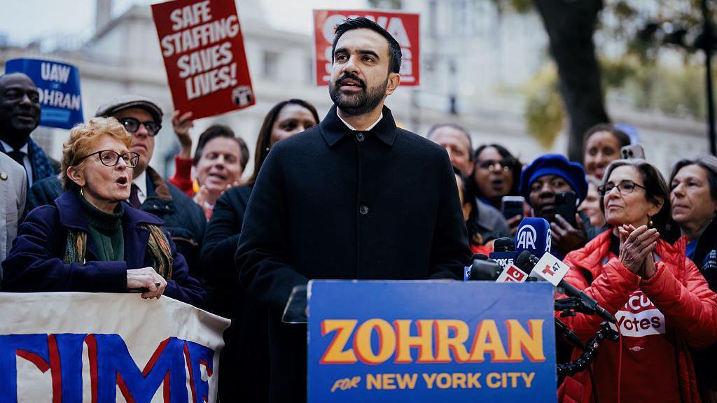 Mamdani speaking at a podium with supporters behind him