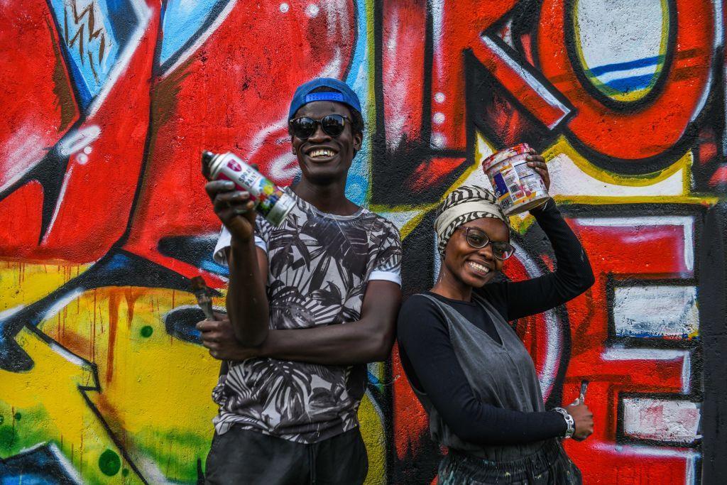 A smiling man and woman stand next to a painted wall, carrying their cans of paint.