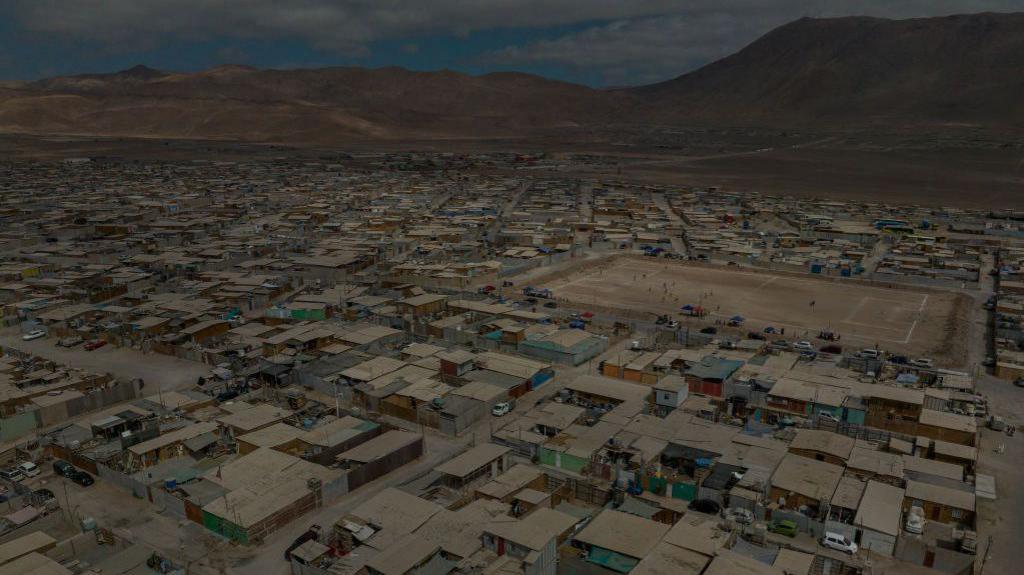 An aerial image showing a dense collection of homes and a dirt playing field in the Flor de Poblacion encampment in Alto Hospicio, Chile. The ground looks dry and dusty and there are barren mountains in the background  