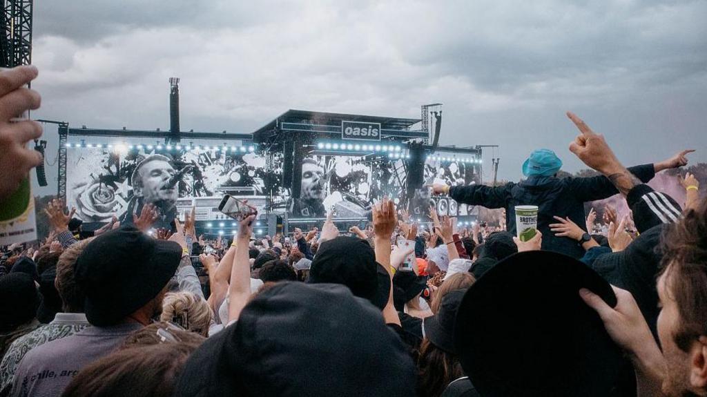Crowd at Oasis gig. Oasis can be seen on the screens beside the stage ahead. 