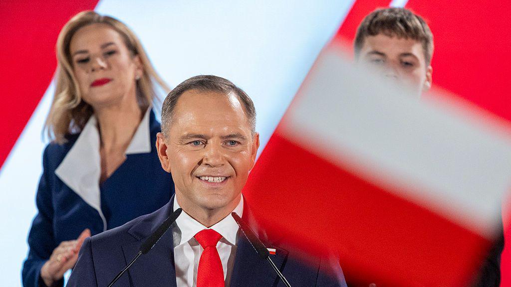 Karol Nawrocki stands smiling at a podium, family members behind him, wearing a dark suit, white shirt and red tie. A Polish flag is out of focus in the foreground.