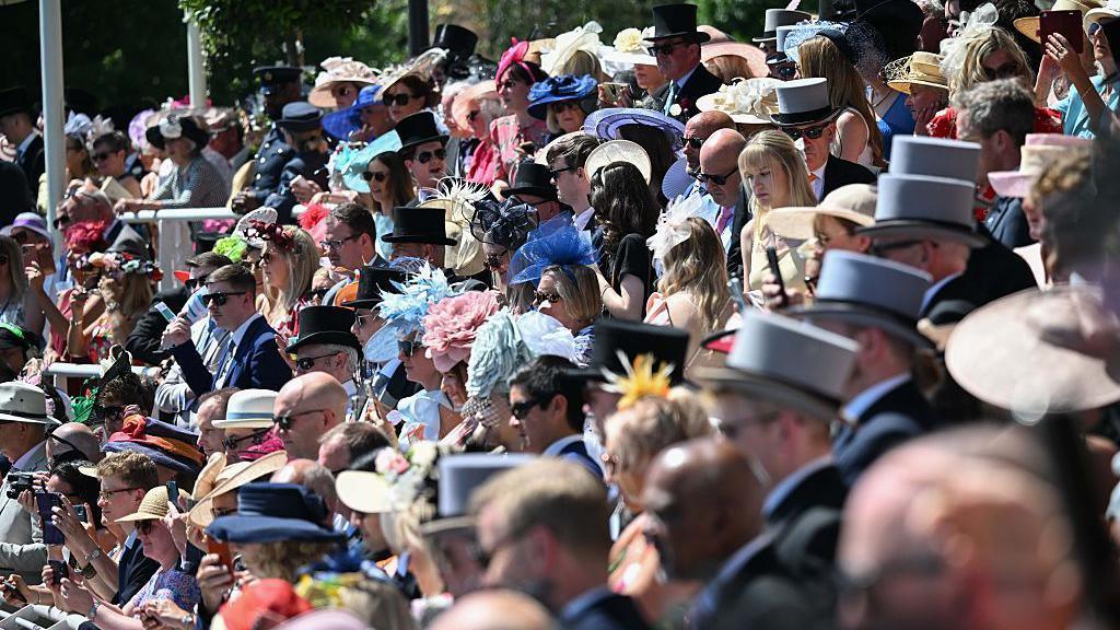 A crowd of people in smart clothing including extravagant hats, sitting on raked seating. It's a sunny day.