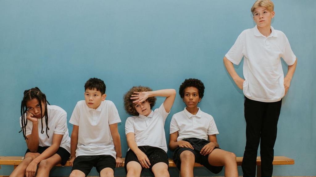 Five young teenage boys sit on a bench in a school gymnasium, looking very hot.