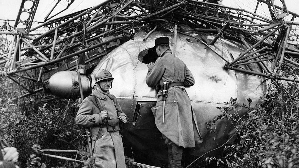 Two French soldiers, in uniforms, with hats and coats, at the scene of the crash of the British airship R101 in the French village of Beauvais. There is debris all around them, and part of the airship. One is looking towards the camera, the other to the ship. 