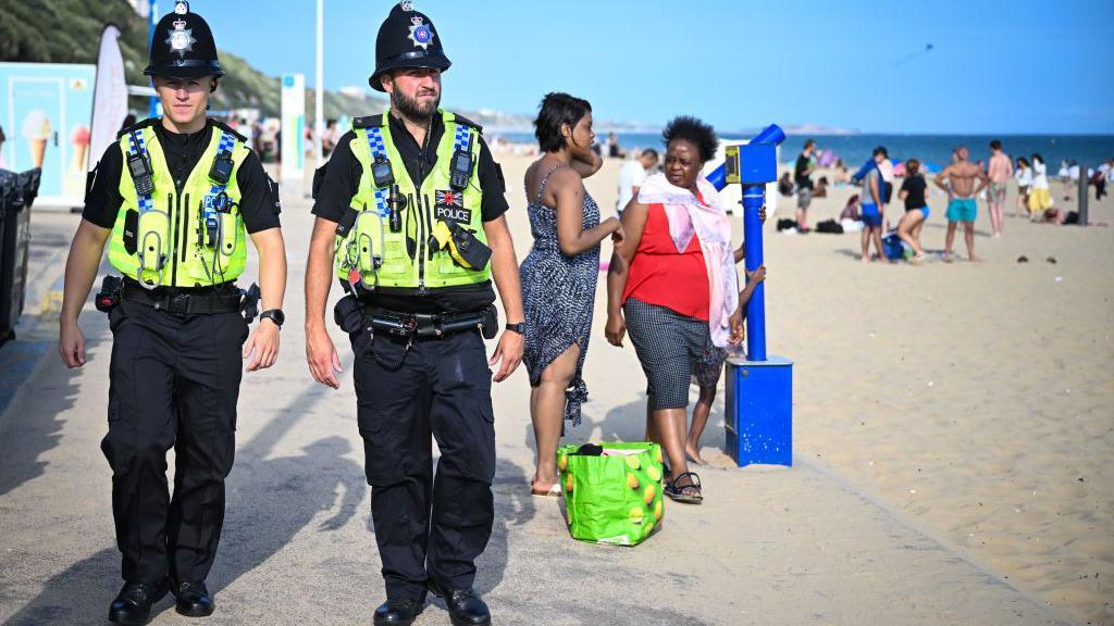 Two white policemen in uniform patrol an esplanade beside Bournemouth beach. Two black women stand behind them.