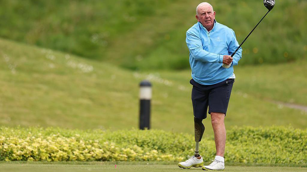 Richie Willis, dyn gwallt gwyn mewn top glas a shorts du, yn chwarae golff - coes brosthetig yw ei goes dde