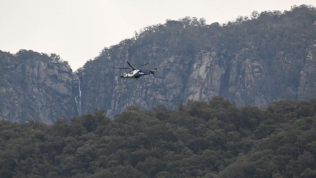 A police helicopter flies over tree tops with a cliff face in the background