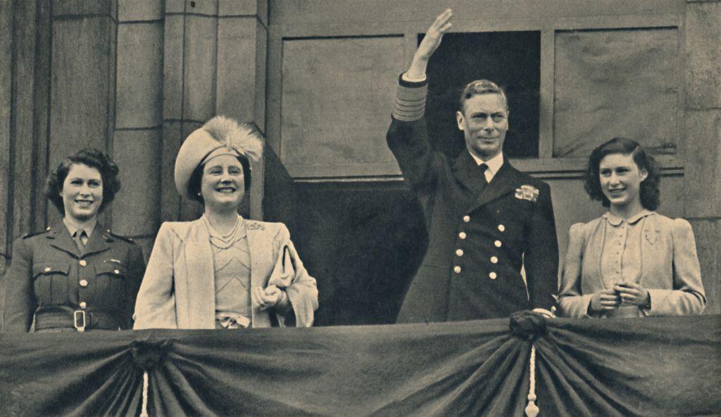 The King, Queen and Princesses Elizabeth and Margaret are smiling from the Royal Balcony at Buckingham Palace. King George VI and Princess Elizabeth are wearing military uniform. The Queen and Princess Margaret are dressed formally.