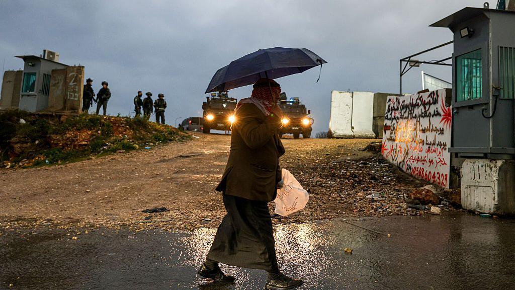 A man walks through a checkpoint, holding an umbrella. 