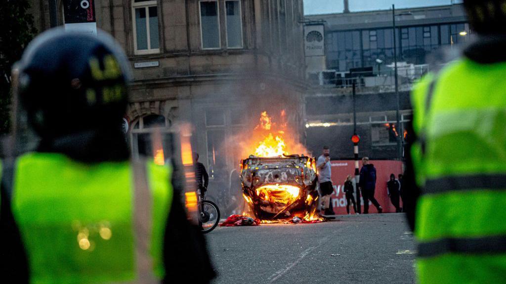 Two police officers stand at a distance from an upturned car on fire.