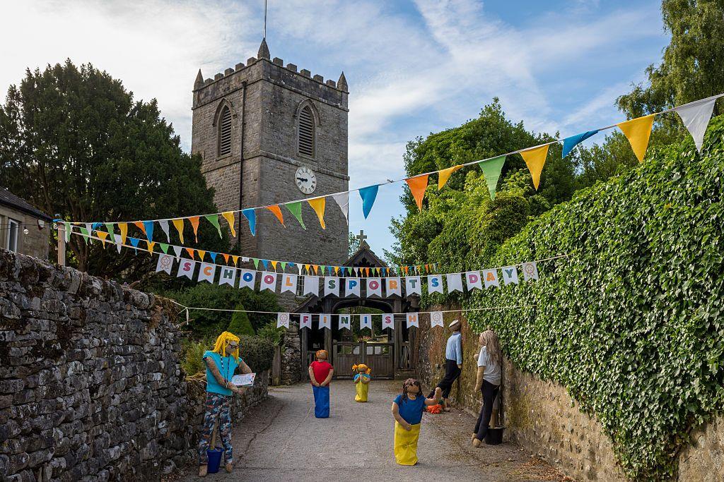 Six scarecrows as seen in this display in front of a church. There is bunting hung over the road and three of the scarecrows are seen taking part in a sack race. The other three are watching on.