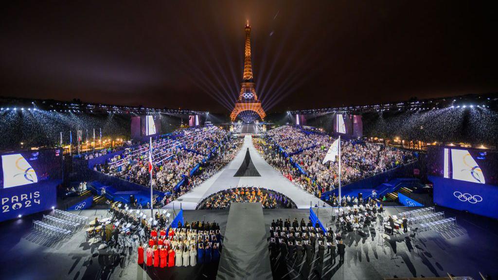 Athletes in front of the Eiffel Tower lit up at night with Olympic branding at either side of hundreds of people